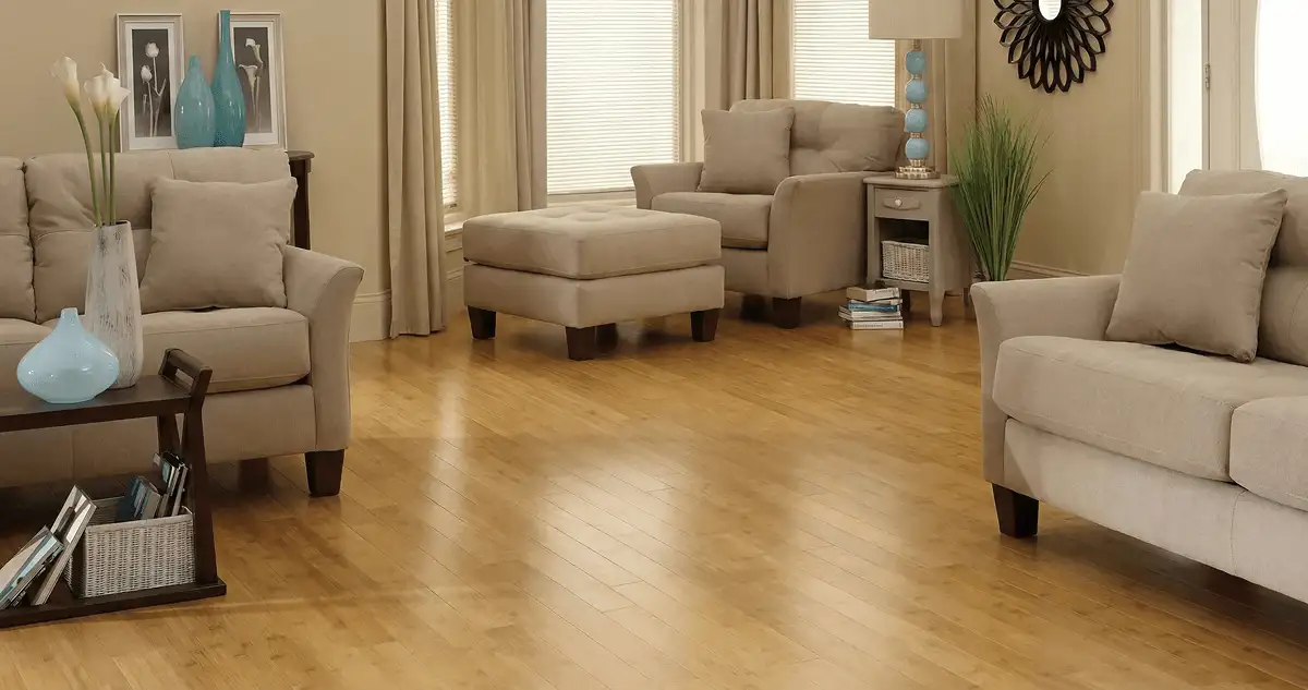 A living room with professionally installed bamboo flooring, beige sofas, a matching ottoman, a side table with a blue lamp, vases, framed pictures, and large windows with blinds and curtains in Arvada, CO